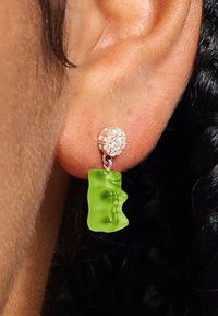 Grüne Ohrringe in Form von Gummibärchen, die mit einem Strassstein-Niet und silberfarbenen Beschlägen versehen sind, und ein verspieltes Design sowie lebendige Farben hervorheben.