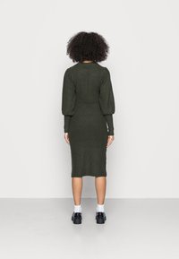 Robe en maille vert olive avec une silhouette ajustée, des manches bouffantes, une texture côtelée et une longueur au genou, associée à des chaussures noires et des chaussettes blanches.