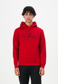Jaunas vyras stovi, apsirengęs raudona džemperio su "Tom Tailor" logotipu ir juodomis kelnėmis, prieš paprastą baltą foną.