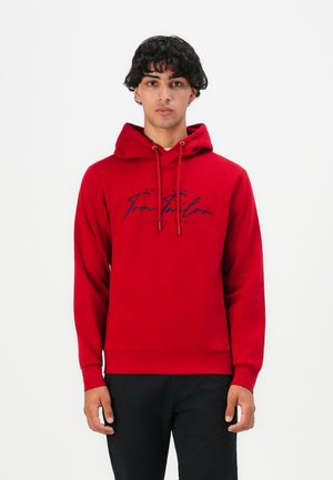 Junger Mann steht, trägt einen roten Kapuzenpullover mit "Tom Tailor"-Logo und schwarze Hosen, vor einem schlichten weißen Hintergrund.