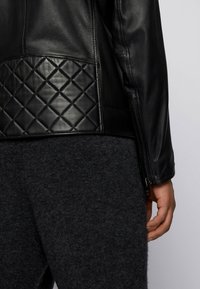 Černá kožená bunda s prošívanou texturou na spodní části, se zipovou kapsou na boku a hladkým povrchem na rukávech.