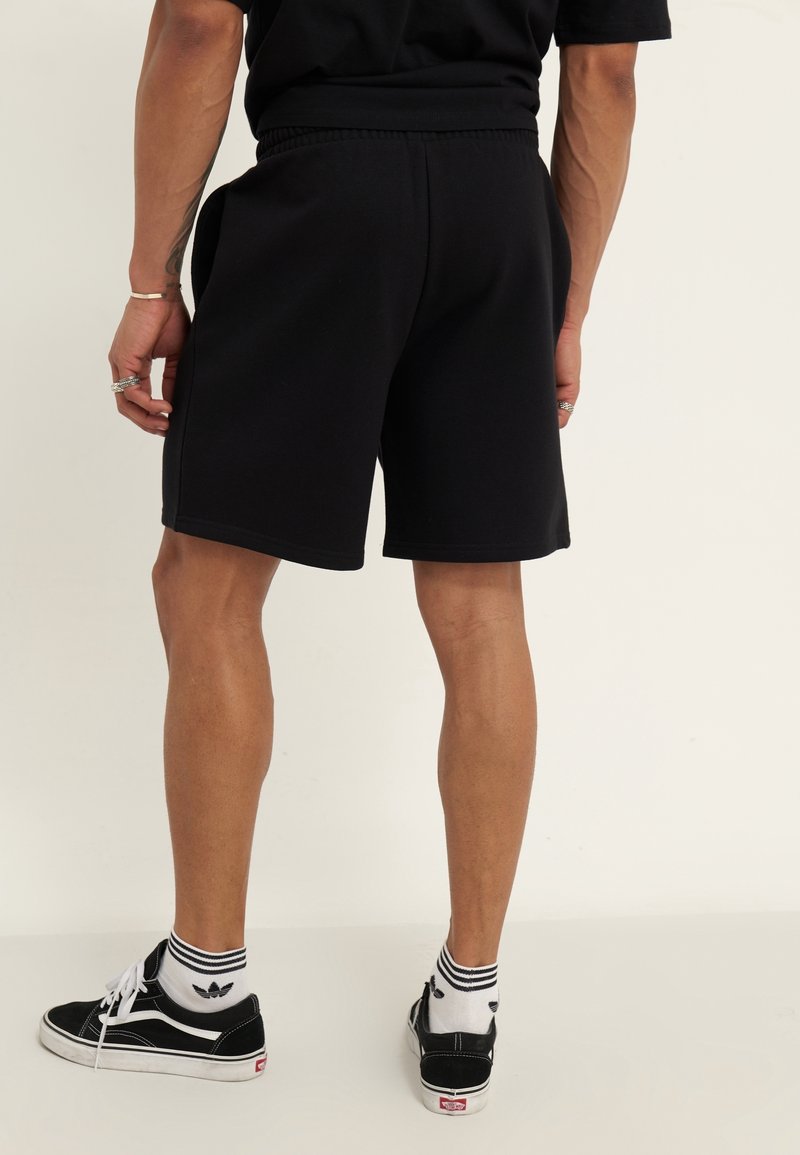 Black cotton shorts with an elastic waistband, straight cut, and side pockets. Paired with black sneakers and white striped socks.
