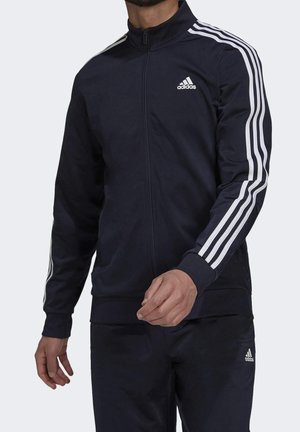 Man draagt een zwarte Adidas trainingsjas met witte strepen op de mouwen en het Adidas-logo op de borst en broek.