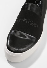 Gros plan sur une sneaker noire slip-on avec bout brillant, semelle blanche et large bande élastique noire marquée "Calvin Klein."