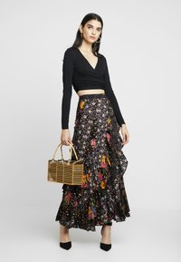 Top preto de manga longa envolvente, combinado com uma saia maxi com padrão floral e babados, adornada com flores multicoloridas. A segurar uma mala de estilo bambu.
