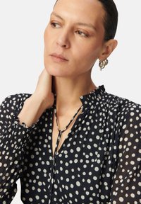 Blusa de lunares en blanco y negro con mangas largas, puños fruncidos y escote en V con detalle de lazo. Tejido ligero y texturizado.