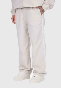 Cilvēks, kura mugurā ir gaiši bežīgas sporta bikses ar baltām sānu svītrām un baltas kedas, stāvot ar vienu roku kabatā.