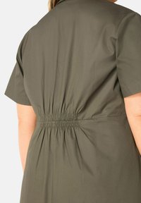 Vestito verde oliva a maniche corte con dettaglio arricciato sul retro, texture liscia e vita aderente, realizzato in un tessuto leggero.