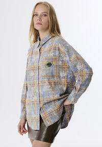 Camicia a quadri nei toni del blu, giallo e marrone, con una tasca frontale decorata con una toppa a forma di pianeta nera e gialla. Tessuto morbido e leggero.