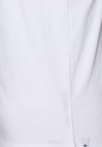 T-shirt bianco in cotone con una texture liscia, vestibilità regolare, cucitura laterale e un piccolo logo verde di coccodrillo vicino all'orlo.