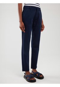 Temno modre slim-fit hlače z stranskimi žepi, v kombinaciji s temnimi sandalmi s prečnimi trakovi in debelim črnim podplatom.