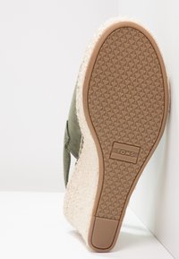 Chaussures vert olive avec un dessus en tissu texturé, semelle intermédiaire en jute et semelle extérieure en caoutchouc avec une adhérence à motif. Logo "TOMS" sur la semelle.