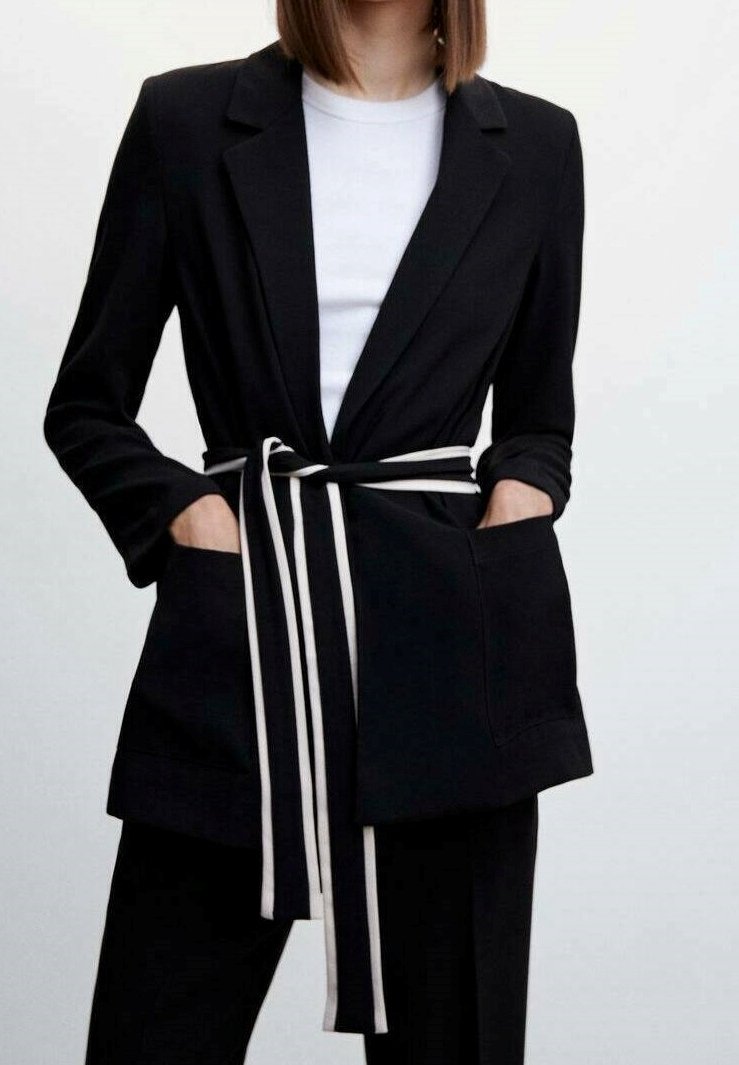 Femme portant un blazer noir avec de grandes poches et une ceinture rayée noire et blanche sur une chemise blanche.