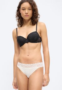 Ženska s kodrastimi lasmi, oblečena v črni čipkasti modrček in belo spodnje perilo z čipko ter logotipom Tommy Hilfiger na pasu, stoji na belem ozadju.