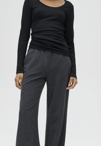 Persona che indossa una maglia nera a maniche lunghe e pantaloni larghi grigio scuro, in piedi davanti a uno sfondo semplice.