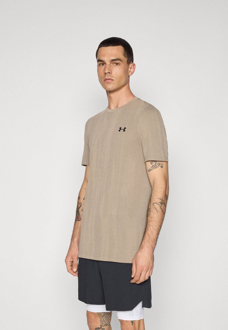 Beige t-skjorte med korte ermer til trening med et teksturert mønster, med en svart logo på øvre venstre bryst. Kombinert med mørke shorts.