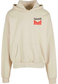 Cremefarget hettegenser med kengurulomme, elastisk midje og rød "UpscaleSt" logo patch på brystet. Myk tekstur.