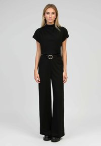 Jumpsuit noir avec un col montant, des manches courtes, une coupe large, taille cintrée avec une ceinture à boucle circulaire, texture de tissu lisse.