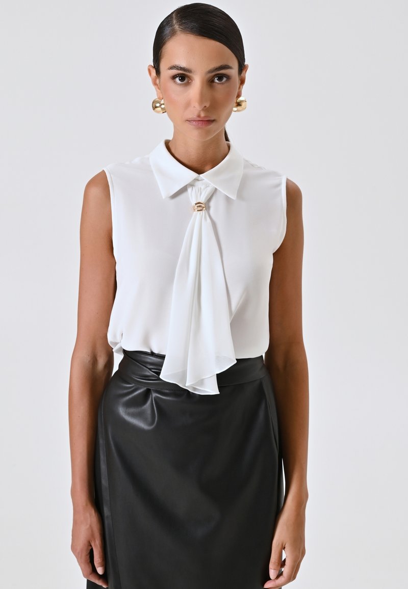Vrouw met naar achteren gekamd haar, gouden oorbellen, een mouwloze witte blouse met geplooid voorkantdetail en een hooggesneden zwarte leren rok.