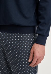 Navy katoenen top met geribbelde manchetten, gecombineerd met donkergekleurde onderkleding met lichte blauwe diamantvormen. Close-up van een hand op de stof.