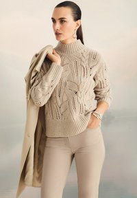 Frau trägt einen beigen Kabelstrickpullover und Hose, hält einen passenden Mantel über einer Schulter, mit silbernen Ohrringe und Armband.