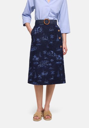 Mujer vistiendo una falda midi azul con botones y estampado costero, una blusa azul y sandalias beige de tacón, de pie con las manos en los bolsillos.