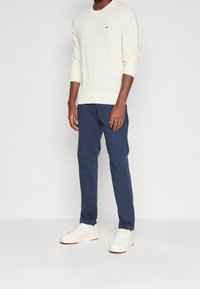 Man in een gebroken witte lange mouwen trui, marineblauwe rechte broek en gebroken witte sneakers tegen een effen lichte achtergrond.