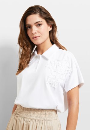 White polo shirt with a textured floral design on the left side and short sleeves, paired with high-waisted beige pants.