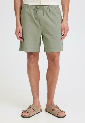 Homme portant un short vert clair à cordon de serrage et des sandales en daim beige, debout devant un fond clair uni.