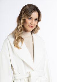 Kremowy szlafrok o miękkiej fakturze, z szerokimi klapami i paskiem w talii, noszony na jasnobeżowej bluzce z wysokim kołnierzem.