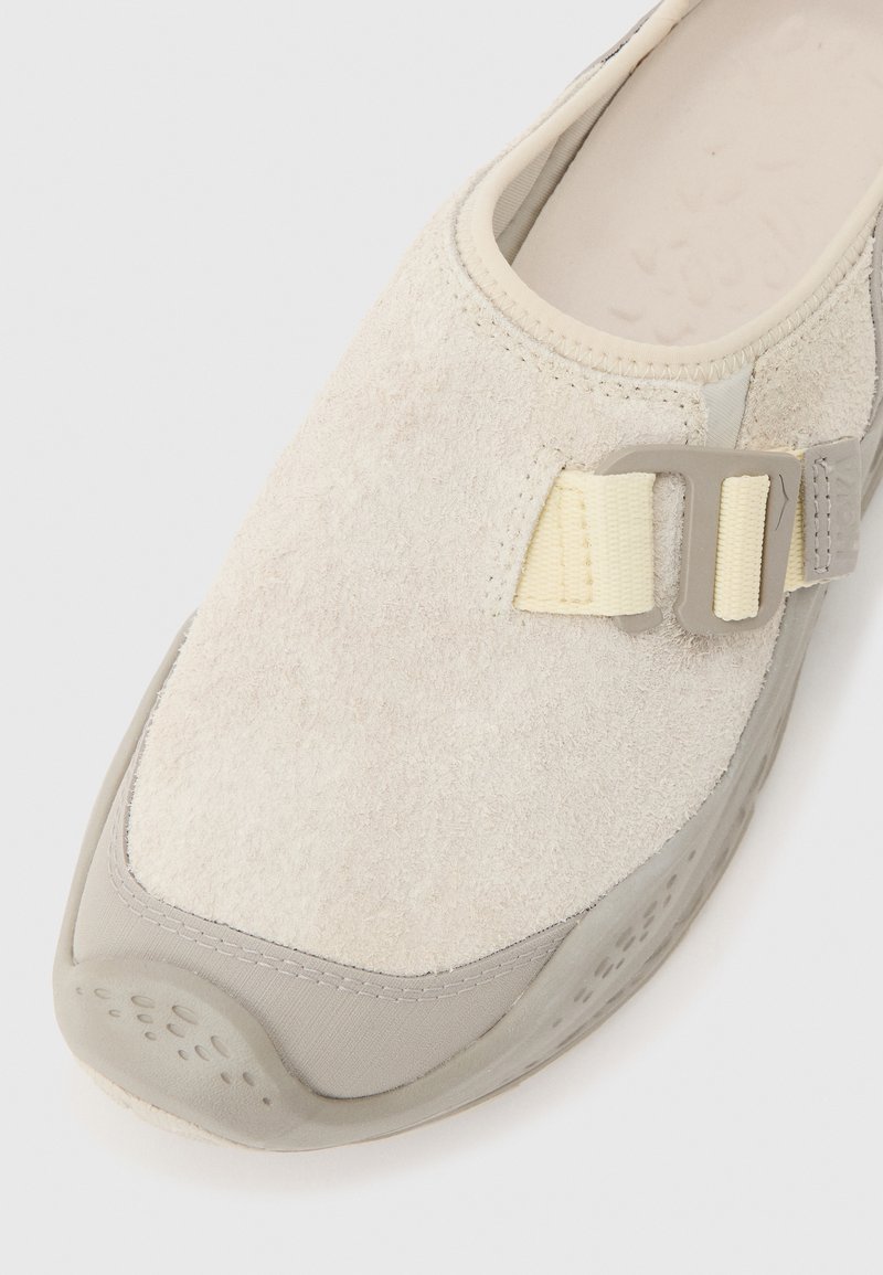 Beige instapper met een gestructureerd bovenwerk, een rubberen zool en een verstelbare crèmekleurige band die wordt vastgehouden door een grijs kunststof gesp.