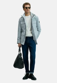 Man wearing light gray jacket, white sweater, dark blue jeans, black shoes, sunglasses, holding black leather duffel bag with one hand.