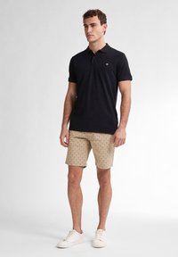 Polo nero a maniche corte, abbinato a pantaloni corti beige chiaro con un motivo geometrico e sneakers bianche.