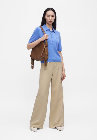 Femme portant un polo bleu à manches courtes et un pantalon beige à jambes larges, tenant un sac à bandoulière en cuir marron avec des pompons.
