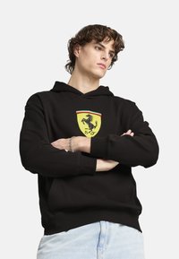 Sort hættetrøje med et gult Ferrari-logo på brystet, med grøn og rød detaljering. Blanding af bomuldsstof, afslappet pasform.