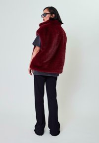 Bordo dirbtinio kailio liemenė su aukštu kaklu, derinama su tamsiu tekstūruotu marškinėliu ir smailiomis kelnių juostelėmis, turinčiomis išlaikytas apatinės dalies dalis.