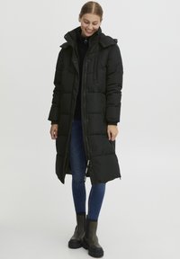 Svart dunjakke med lang design, glidelås og trykknapplukking, sidelommer og avtakbar hette. Brukt over mørkeblå jeans og støvler.