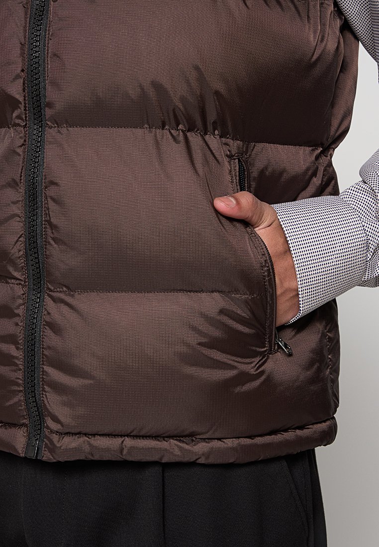Gilet marron rembourré avec une finition texturée, fermeture éclair à l'avant et poches latérales. Porté par-dessus une chemise à motifs, avec une ceinture noire visible.