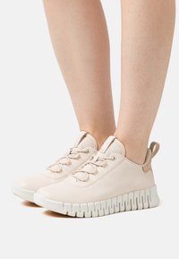 Fötter i beige sneakers med texturerade mesh-tungor och tjocka vita sulor med zigzagmönster mot en vit bakgrund.