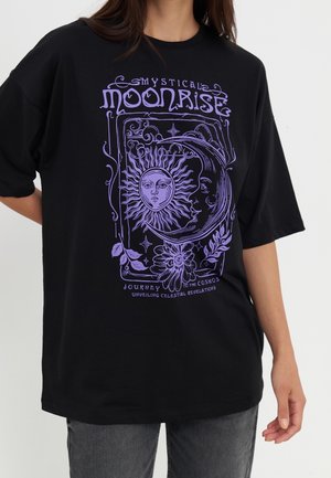 Person iført en sort oversized t-shirt med lilla mystisk sol- og måne-grafik og teksten "Mystisk måneopgang rejse til kosmos."