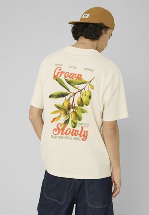 Jeune personne portant un t-shirt beige avec une branche d'olivier et le texte « Grown Slowly Tradition Press Gently », associé à un jean foncé et une casquette moutarde.