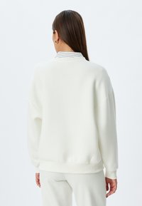 Felpa bianca oversized con polsini e orlo a costine, caratterizzata da una leggera trama. Design con colletto sovrapposto a una camicia a righe.