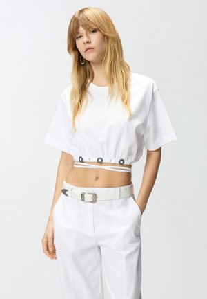 Jeune femme aux longs cheveux blonds porte un t-shirt court blanc avec des boutons décoratifs et un pantalon blanc assorti avec ceinture, debout avec une main dans la poche.