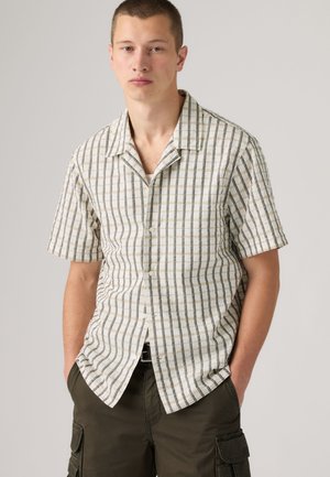 Jeune homme portant une chemise crème à manches courtes avec des rayures verticales et horizontales, associée à un short cargo foncé, debout les mains dans les poches.