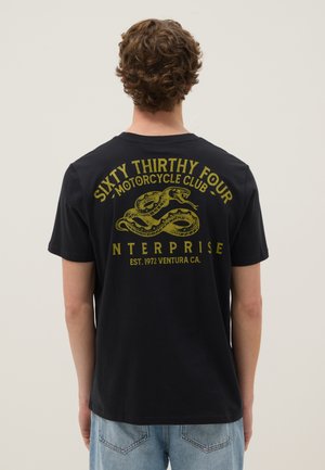 Persoon met krullend bruin haar die een zwart T-shirt draagt met een gele slang en de tekst "Sixty Thirty Four Motorcycle Club" op de rug.