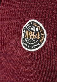 Bordeaux gestrickter Pulloverstoff mit einem runden schwarzen Patch, das weißen und goldenen Text sowie ein Globusdesign zeigt; detaillierte Textur sichtbar.