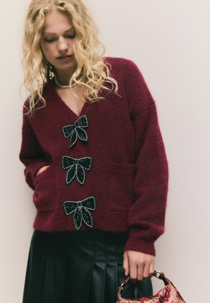 Burgundy wool cardigan with a V-neck, featuring three black beaded bow accents. Model wears a black pleated leather skirt and holds a floral handbag.