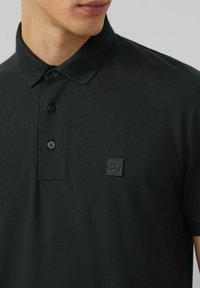 Schwarzes Poloshirt aus strukturiertem Stoff, mit einer Drei-Knopf-Blende und einem kleinen, quadratischen Logo-Patch auf der Brust.