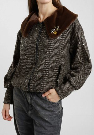 Donna che indossa un giubbotto marrone a spina di pesce con paillettes, colletto in ecopelliccia marrone, spilla con gioielli, zip nera e jeans neri lavati con una mano in tasca.