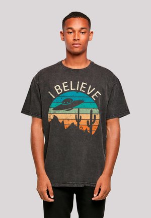 Black graphic t-shirt featuring an "I believe" design with an alien spaceship, mountains, and cacti in sunset colors. Short sleeves, loose fit.